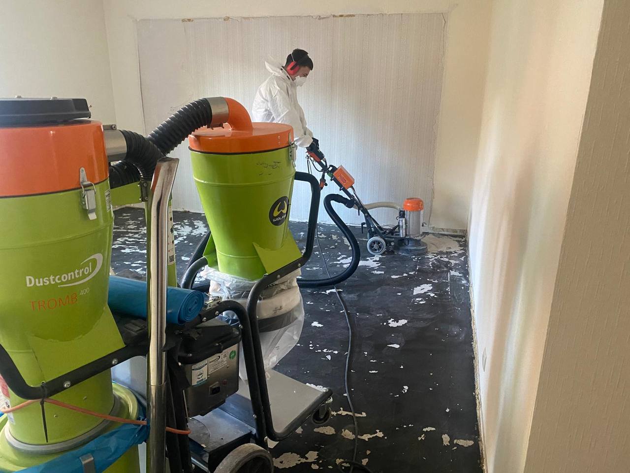 Fachkraft im Schutzanzug entfernt asbesthaltigen Bodenbelag mit Schleifmaschine und Industrieentstauber